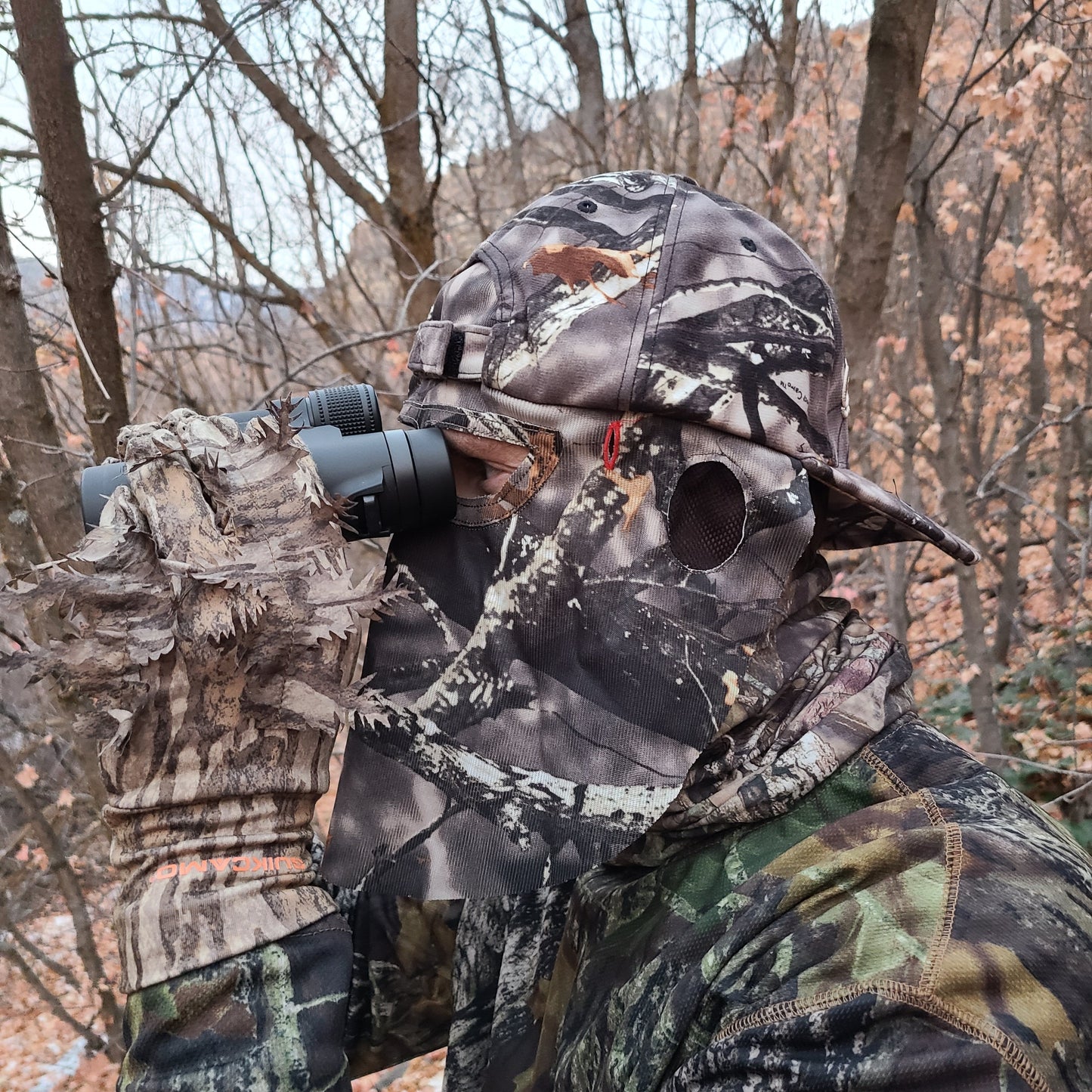 Man Using Binoculars while Wearing a QuikCamo Mathews Lost Camo Hat Backwards with Face Mask. There is a forest or woods in the background
